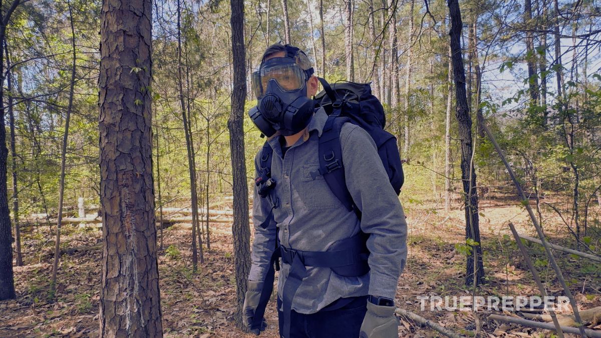 A man walking through the woods wearing a hiking backpack, gloves, and gas mask.